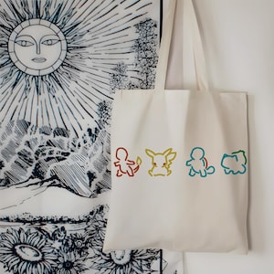May include: Off-white tote bag featuring colorful line art of four cartoon characters. The bag hangs against a backdrop of a black and white sun and floral tapestry. The cartoon characters are outlined in brown, yellow, blue, and green.