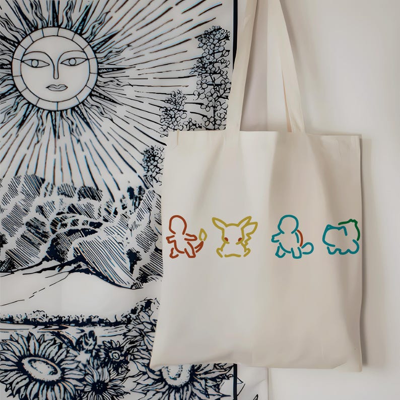 May include: Off-white tote bag featuring colorful line art of four cartoon characters. The bag hangs against a backdrop of a black and white sun and floral tapestry. The cartoon characters are outlined in brown, yellow, blue, and green.