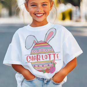 May include: White t-shirt with a colourful Easter egg design, bunny ears, and the name "CHARLOTTE" in pink. The egg has horizontal stripes and is decorated with flowers. A children's top.