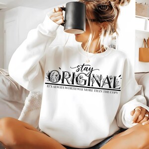 May include: White sweatshirt with the words "stay ORIGINAL" in black script and block letters, with floral accents. The text "IT'S ALWAYS WORTH WAY MORE THAN THE COPY" is below. A person is holding a black mug.