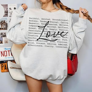 Puede incluir: Una sudadera de cuello redondo gris claro con un bloque de texto negro que enumera varios adjetivos positivos como Faithful, Devoted y Unconditional, con la palabra Love escrita en una letra cursiva negra grande superpuesta al texto.
