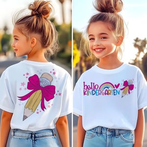 May include: A white t-shirt featuring a graphic of a yellow pencil with a large magenta bow and small pink stars. The front design includes the text "hello KINDERGARTEN" in colourful letters with a small rainbow and a heart icon above the word.