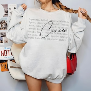 May include: A light gray sweatshirt with the word "Cancer" in a handwritten font, accompanied by a list of descriptive words. The sweatshirt has long sleeves and a crew neck.