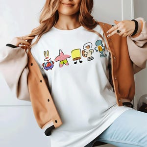 Puede incluir: Una camiseta blanca con un estampado gráfico minimalista de cinco personajes de dibujos animados en fila, incluyendo un cangrejo rojo, una estrella de mar rosa, una esponja amarilla, una ardilla blanca y un pulpo azul verdoso, bajo una chaqueta.