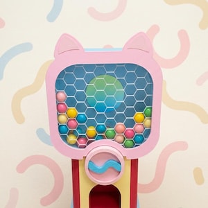 Puede incluir: Una máquina de chicles rosa con detalles de orejas de gato, llena de chicles de colores. La máquina tiene una ventana de rejilla hexagonal y una base azul y amarilla. El fondo presenta un patrón pastel.