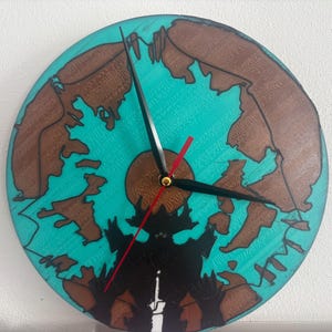 May include: Round wall clock with a teal background and brown landmass design. The clock features black hands, a red second hand, and a brown sun-like center. The clock's design includes a black silhouette at the bottom.