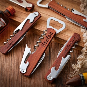 Könnte beinhalten: Mehrere Weinöffner-Werkzeuge aus Holz mit Akzenten aus Edelstahl werden auf einer Holzoberfläche präsentiert. Jedes Werkzeug hat einen Korkenzieher, einen Flaschenöffner und ein Messer. Einige Werkzeuge sind mit Namen graviert: Lenni, Stefan und Clarissa. Eine Flasche Cointreau ist sichtbar.