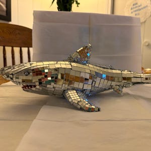 Könnte beinhalten: Eine dekorative Hai-Figur, die mit kleinen, reflektierenden Spiegelfliesen bedeckt ist. Der Hai hat eine Rückenflosse, Brustflossen und eine Schwanzflosse. Das Maul ist geöffnet und zeigt rote Zähne. Die Figur wird auf einer weißen Oberfläche präsentiert.