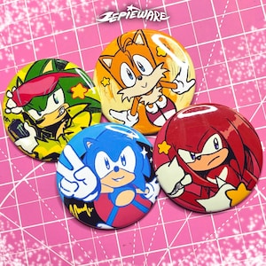 May include: A collection of five round pinback buttons featuring characters from the Sonic the Hedgehog franchise. The buttons showcase Sonic, Tails, Knuckles, and other characters in vibrant colors. The top of the image has the text "ZEPIEWARE".