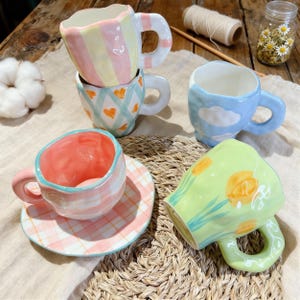 May include: A collection of colorful ceramic mugs with unique designs. One mug features vertical pastel stripes, another has blue hearts, and a third has a cloud design. A fourth mug has a floral pattern, and a fifth has a pink and white plaid pattern.