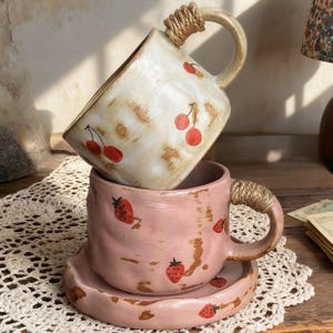 Puede incluir: Dos tazas de cerámica, una color crema con diseños de cerezas rojas y otra rosa con diseños de fresas, están apiladas. Ambas tazas tienen asas envueltas con cuerda. La taza rosa se asienta sobre un platillo a juego, creando una estética rústica encantadora.
