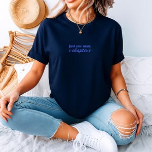 Può includere: T-shirt blu navy con la scritta "Just one more chapter" in blu chiaro. La maglietta è a maniche corte e di stile casual. Il testo è centrato sul petto. È abbinata a jeans azzurri e scarpe da ginnastica bianche.
