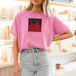 May include: A pink t-shirt with a graphic design. The design features a red silhouette of a person with a sword, mountains, and the text "Girl just wanna read". The shirt is paired with light blue jeans.