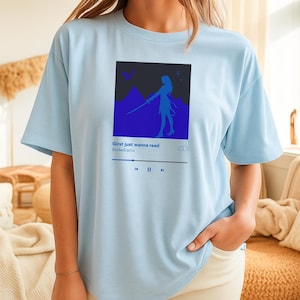 May include: Pale blue t-shirt with a blue graphic of a person with a sword, mountains, and the text "Girlst just wanna read." The design is in a music player style. The brand name "BookedUpCo" is also visible.