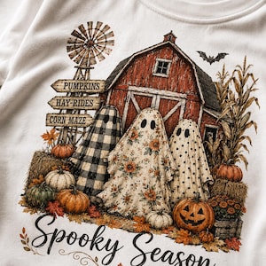 May include: A cream-colored sweatshirt featuring a graphic of three ghosts in front of a red barn, a windmill, and corn stalks. The ghosts wear buffalo plaid, floral, and polka dot patterns. Text reads PUMPKINS, HAY-RIDES, CORN MAZE, and Spooky Season.