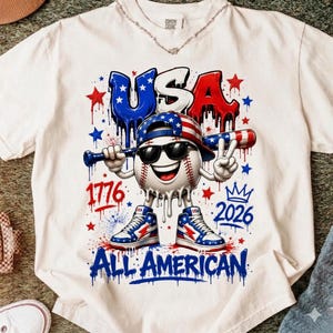 May include: White t-shirt featuring a cartoon baseball character wearing sunglasses, a baseball cap, and sneakers, holding a bat and making a peace sign. The shirt has the word "USA" and "ALL AMERICAN" in blue and red, with the numbers "1776" and "2026".