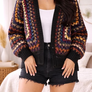 Peut inclure: Un cardigan en crochet multicolore avec une bordure noire et un col en V. Le cardigan présente un mélange de couleurs, dont le bleu, le jaune, le rouge et le marron. Le modèle porte un short en jean noir et un haut blanc.