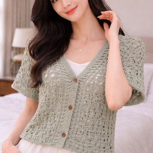 Pode incluir: Um cardigan de crochê verde claro de manga curta, com decote em V e botões de madeira. O design de tecido aberto revela uma blusa branca por baixo. O cardigan é de uma cor verde suave e discreta, ideal para sobrepor.
