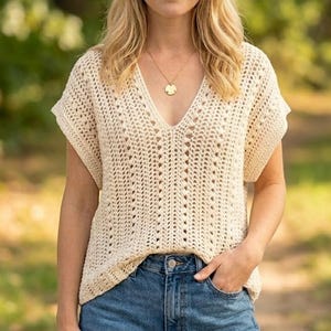 May include: A cream-coloured, short-sleeved crochet top with a deep V-neck. The top features an open, textured pattern and is worn with blue denim jeans. A gold-coloured necklace with a circular pendant is visible.