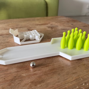 Op de afbeelding: Een miniatuur bowlingspelset met een witte plastic baan, een zilveren bal en tien neon groene bowlingkegels. Een wit plastic stuk houdt de kegels vast. De set staat op een houten tafel.