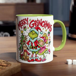 Könnte beinhalten: Ein weißer Keramikbecher mit hellgrünem Henkel und Innenraum, verziert mit einer bunten Illustration einer grünen Kreatur im Weihnachtsmannkostüm, einem jungen Mädchen und einem Hund, mit dem roten Schriftzug Merry Grinchmas darüber.