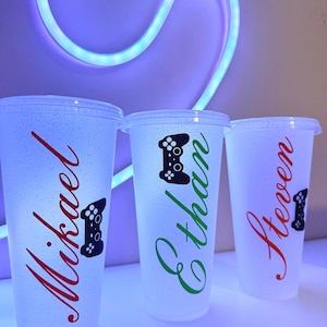 Puede incluir: Tres vasos de plástico transparente con tapas, cada uno personalizado con un nombre en un color diferente: Mikael en rojo, Ethan en verde y Steven en rojo. Cada vaso presenta un gráfico de mando de videojuegos negro.