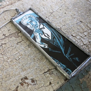 May include: A rectangular pendant necklace featuring a black and blue illustration of a man in a suit. The pendant is framed in silver metal and hangs from a silver chain. The image has a vintage comic book style.