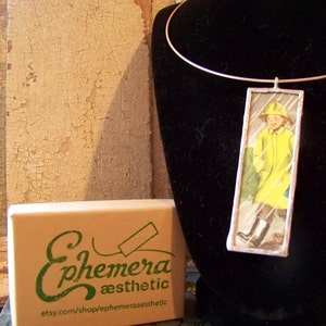 Puede incluir: Un collar de plata con un colgante rectangular de vidrio que presenta una ilustraci&oacute;n vintage de un ni&ntilde;o con un impermeable amarillo. El colgante est&aacute; enmarcado con un borde plateado. El collar se muestra en un busto de maniqu&iacute; de terciopelo negro. El texto "Ephemera aesthetic" y "etsy.com/shop/ephemeraaesthetic" est&aacute; impreso en una caja marr&oacute;n.