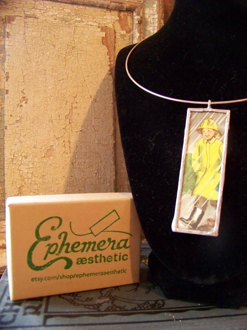 Puede incluir: Un collar de plata con un colgante rectangular de vidrio que presenta una ilustraci&oacute;n vintage de un ni&ntilde;o con un impermeable amarillo. El colgante est&aacute; enmarcado con un borde plateado. El collar se muestra en un busto de maniqu&iacute; de terciopelo negro. El texto "Ephemera aesthetic" y "etsy.com/shop/ephemeraaesthetic" est&aacute; impreso en una caja marr&oacute;n.