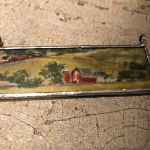 Peut inclure: Un pendentif rectangulaire en verre teinté représentant une scène de campagne avec une grange rouge, des arbres verts et un chemin sinueux. Le pendentif est doté d'un cadre argenté et est parfait pour ajouter une touche de charme rustique à n'importe quel espace.