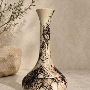 May include: A tall, slender vase with a flared rim and a rounded base, featuring a cream-colored surface decorated with an intricate, dark black marbled pattern that extends from the bottom up to the narrow neck of the vessel.