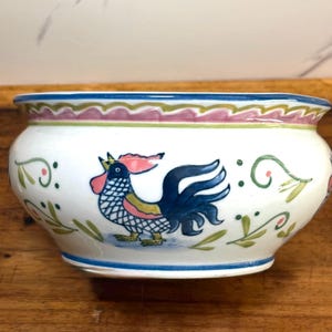 May include: White ceramic bowl with a hand-painted rooster design. The bowl features a blue rim, a pink and green decorative border, and green leaf accents. The rooster is depicted in blue, red, yellow, and green.