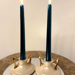 Pode incluir: Duas velas cônicas teal acesas em castiçais creme com alças douradas. Os castiçais estão sobre uma superfície espelhada, refletindo as velas e os suportes. O fundo é de cor neutra.