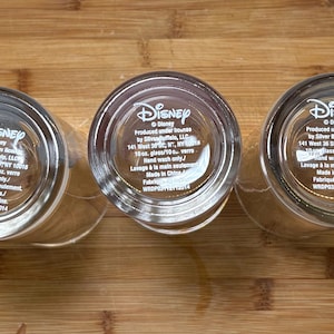 May include: Three clear Disney glasses with the Disney logo and product information printed on the base. Each glass is 29.5 cl and made in China. The glasses are on a wooden surface.