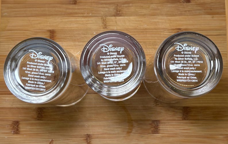 May include: Three clear Disney glasses with the Disney logo and product information printed on the base. Each glass is 29.5 cl and made in China. The glasses are on a wooden surface.