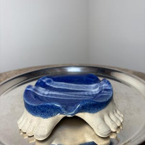 Może przedstawiać: Ceramiczna mydelniczka o unikalnym designie. Mydelniczka ma niebiesko-biały falisty wzór na górze, wsparty na dwóch beżowych stopach. Mydelniczka jest umieszczona na srebrnej tacy.