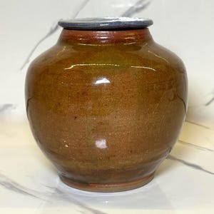 Puede incluir: Un jarrón de cerámica marrón con tapa. El jarrón tiene un cuerpo redondeado y un borde ligeramente ensanchado. La tapa es de un color más oscuro que el jarrón. El jarrón está esmaltado y tiene un acabado brillante. El jarrón mide unos 15 cm de alto y 13 cm de ancho.