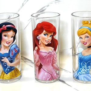 May include: Three clear glass tumblers featuring Disney princesses Snow White, Ariel, and Cinderella. Snow White wears a blue and yellow dress. Ariel is in a pink dress. Cinderella is in a blue dress.