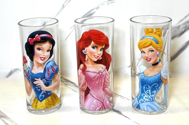 May include: Three clear glass tumblers featuring Disney princesses Snow White, Ariel, and Cinderella. Snow White wears a blue and yellow dress. Ariel is in a pink dress. Cinderella is in a blue dress.