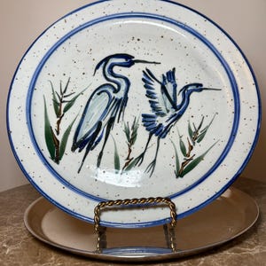 May include: White ceramic plate with a blue rim and a hand-painted design of two blue herons and green reeds. The plate is speckled with brown dots and is displayed on a metal stand.