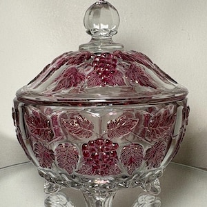May include: A clear glass candy dish with a lid, featuring a grape and leaf design in a deep red color. The dish sits on a pedestal base with three decorative feet. The lid has a round finial.