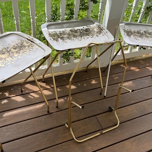 May include: Three vintage metal TV trays with folding legs. The trays have a light gray background with a floral design in shades of gold and brown. The legs are gold and silver. The trays are set up on a wooden deck.