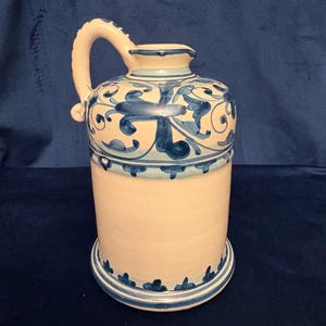 Op de afbeelding: Een keramische kan met een handvat, met een crèmekleurige body en ingewikkelde blauwe bloempatronen. De kan heeft een cilindrische basis en een afgeronde bovenkant met een tuit. Het handvat is gebogen en het ontwerp bevat een blauwe rand.