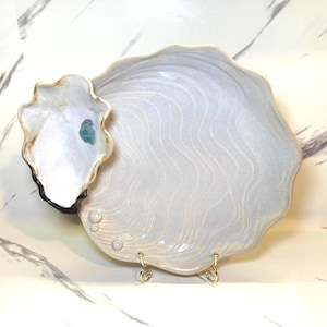 May include: A ceramic oyster-shaped serving dish with a matching small dish attached. The large dish is white with a wavy pattern, and the small dish has a gold rim. The dish is displayed on a gold stand.
