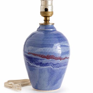 May include: A ceramic table lamp base with a glossy blue glaze featuring horizontal bands of white and dark red. The lamp has a metallic socket at the top and a white electrical cord with a plug extending from the base of the ceramic body.