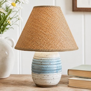 May include: A table lamp featuring a textured, conical lampshade in a natural tan woven material and a rounded, ribbed ceramic base with a speckled glaze in horizontal bands of white and light blue, resting on a flat, circular base.