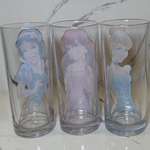 May include: Three clear glass tumblers with Disney princess designs. Each glass features a different princess: Snow White in a blue dress, Ariel in a pink dress, and Cinderella in a blue dress. The glasses are cylindrical with a slightly flared base.