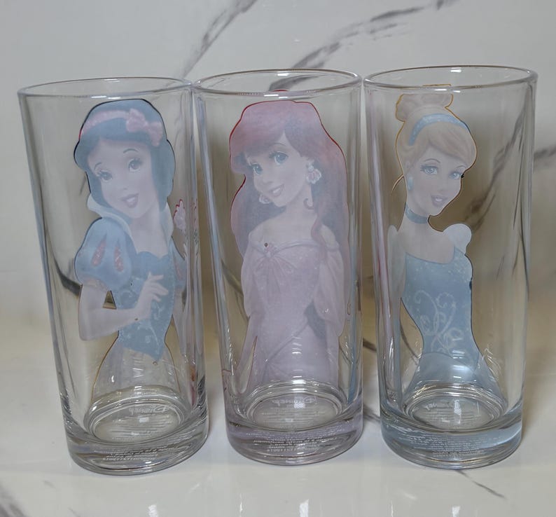 May include: Three clear glass tumblers with Disney princess designs. Each glass features a different princess: Snow White in a blue dress, Ariel in a pink dress, and Cinderella in a blue dress. The glasses are cylindrical with a slightly flared base.