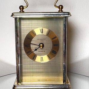 May include: A rectangular, vintage Hamilton Quartz clock with a gold-tone face and Roman numerals. The clock has a silver-tone frame and a gold handle. The words "HAMILTON" and "QUARTZ" are visible on the clock face. The clock is marked "MADE IN GERMANY".