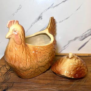 May include: A ceramic hen-shaped container with a lid. The container is a warm brown color with a lighter brown comb and wattle. The lid is a smaller version of the hen's body. The container is sitting on a wooden surface.